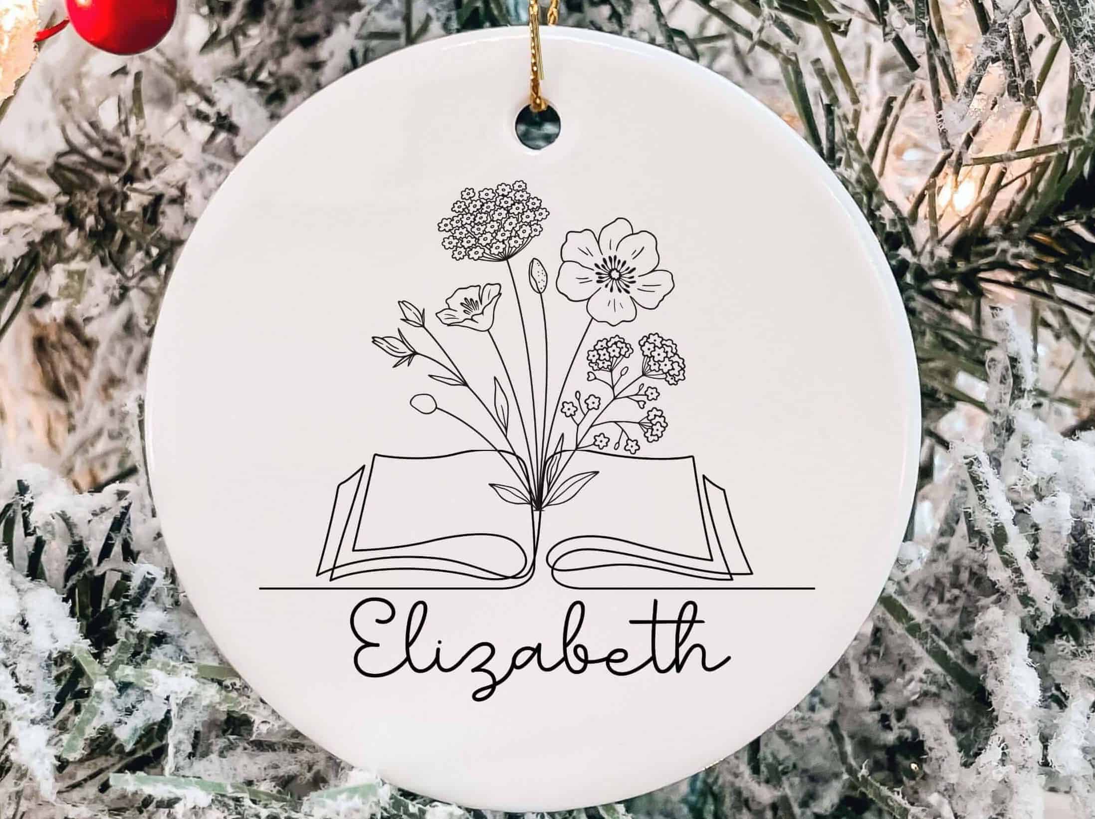 White holiday ornament with a drawing of a book with wildflowers growing out of it, and the name Elizabeth below