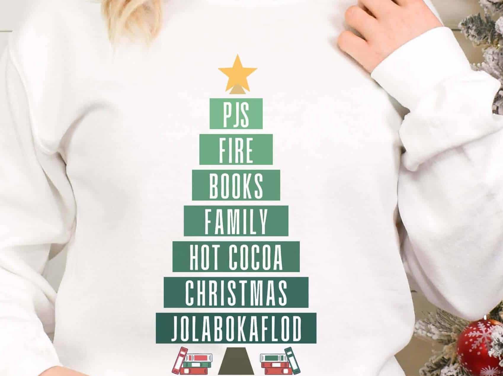 Woman wearing a sweatshirt with layers of a tree reading PJs, Fire, Books, Family, Hot cocoa, Christmas, Jolabokaflod
